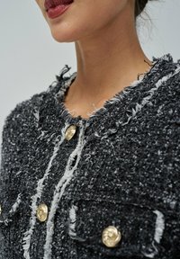 Veste texturée noir et blanc avec des bords effilochés, boutons dorés et deux poches avant. La vue en gros plan met en valeur les détails du col et des boutons.