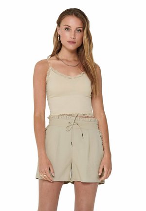Young woman with long hair wearing beige lace-trimmed camisole and high-waisted shorts with drawstring, standing against white background.
