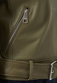 Jachetă din piele verde măslinie cu un fermoar argintiu și detalii cu cataramă, având o vedere de aproape a materialului texturat și neted, precum și a cusăturilor.