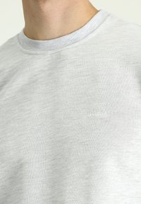 Sudadera gris claro con un cuello redondo acanalado, textura suave y un sutil detalle de logo bordado en el pecho.
