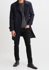 Granatowy płaszcz przeciwdeszczowy z podwójnym zapięciem na guziki, szerokim kołnierzem i długimi rękawami, w połączeniu z szarą koszulką i czarnymi, dopasowanymi spodniami.