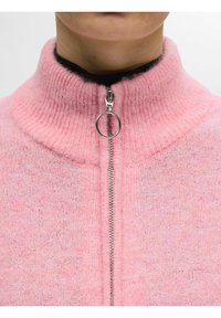 Pull à zip rose flou avec col montant et fermeture éclair argentée. Tissu en maille texturée avec des variations de couleur subtiles.