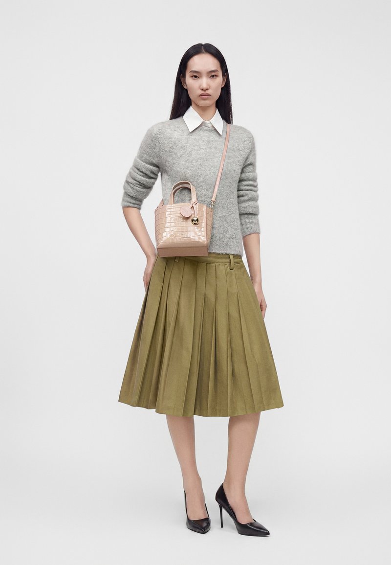 Femme portant un pull gris, une chemise blanche à col, une jupe plissée vert olive, des talons noirs, et portant un petit sac à main beige sur son épaule.