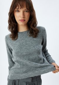Maglioncino grigio lavorato a maglia con scollo rotondo, maniche lunghe e una consistenza morbida, leggermente arricciato all'orlo e indossato sopra pantaloni a vita alta.