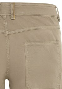 Gros plan de l'arrière d'un pantalon beige montrant une poche, une passants de ceinture avec un détail de couture jaune sur un tissu robuste.