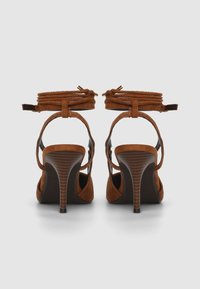 Braune Wildleder-Sandalen mit hohem Absatz, Knöchelriemen, spitzem Zehenbereich und einem strukturierten braunen Absatz. Versehen mit einer gebundenen Detailverzierung am Knöchel.