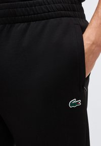 Εγκαρσία λήψη του χεριού ενός ατόμου στην τσέπη μαύρου φλις παντελονιού με ελαστική μέση και πράσινο λογότυπο κροκόδειλου στο μηρό.