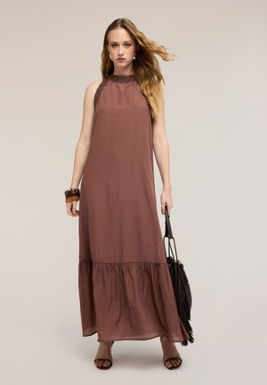 Mujer con vestido largo marrón sin mangas sosteniendo un bolso negro con flecos, llevando pulseras y tacones abiertos, de pie frente a un fondo beige.