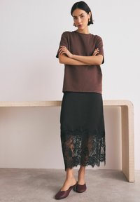 Top corto marrone a maniche corte abbinato a una gonna nera con orlo in pizzo. La modella indossa scarpe scivolate color marsala, in piedi accanto a un tavolo chiaro.