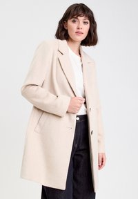 Manteau beige en tissu doux, avec un col en pointe, des manches longues, deux poches avant et une coupe droite qui tombe en dessous des hanches.