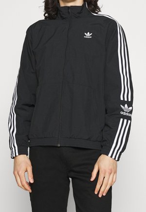 Person trägt eine schwarze Adidas-Trainingsjacke mit weißen Streifen an den Ärmeln sowie dem Adidas-Logo auf der Brust und dem linken Arm.