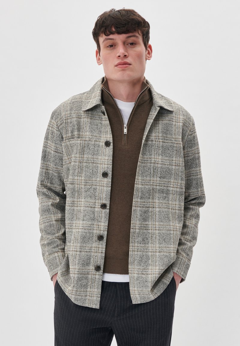 Camisa de cuadros gris de botones para hombre con un acabado texturizado, que cuenta con cuello, bolsillos y un corte relajado sobre un suéter marrón oscuro.