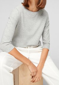 Light grey long-sleeve top with a round neckline and subtle texture, paired with white textured pants. Model seated on a wooden box.