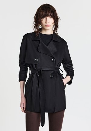 Donna con capelli scuri e bagnati indossa un trench coat nero con cintura sopra un top nero, in piedi con una mano in tasca davanti a uno sfondo neutro.