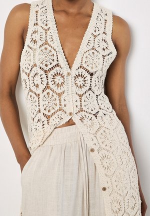 Ärmellose beige gehäkelte Weste mit Blumenmustern und Knöpfen über hellen Leinenhosen mit hoher Taille, eine Hand in der Tasche.