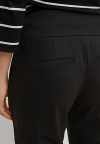 Zwarte op maat gemaakte broek met een gladde textuur, voorzien van twee achterzakken en een tailleband. Gestreepte top zichtbaar boven de tailleband.