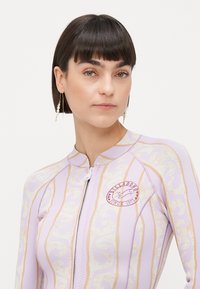 Long-sleeve wetsuit in pastel lavender with gold wave patterns and a front zip. Features a round logo patch on the chest. Smooth texture.