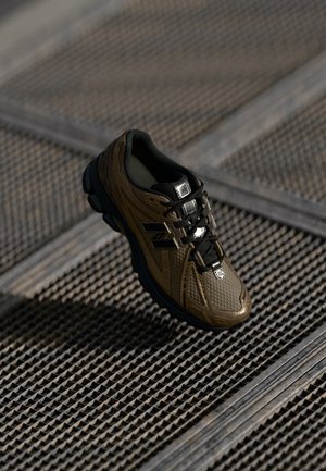 Braun-schwarzer New Balance Sneaker mit strukturiertem Design, der auf einer gemusterten Metalloberfläche mit starken Schatten liegt.