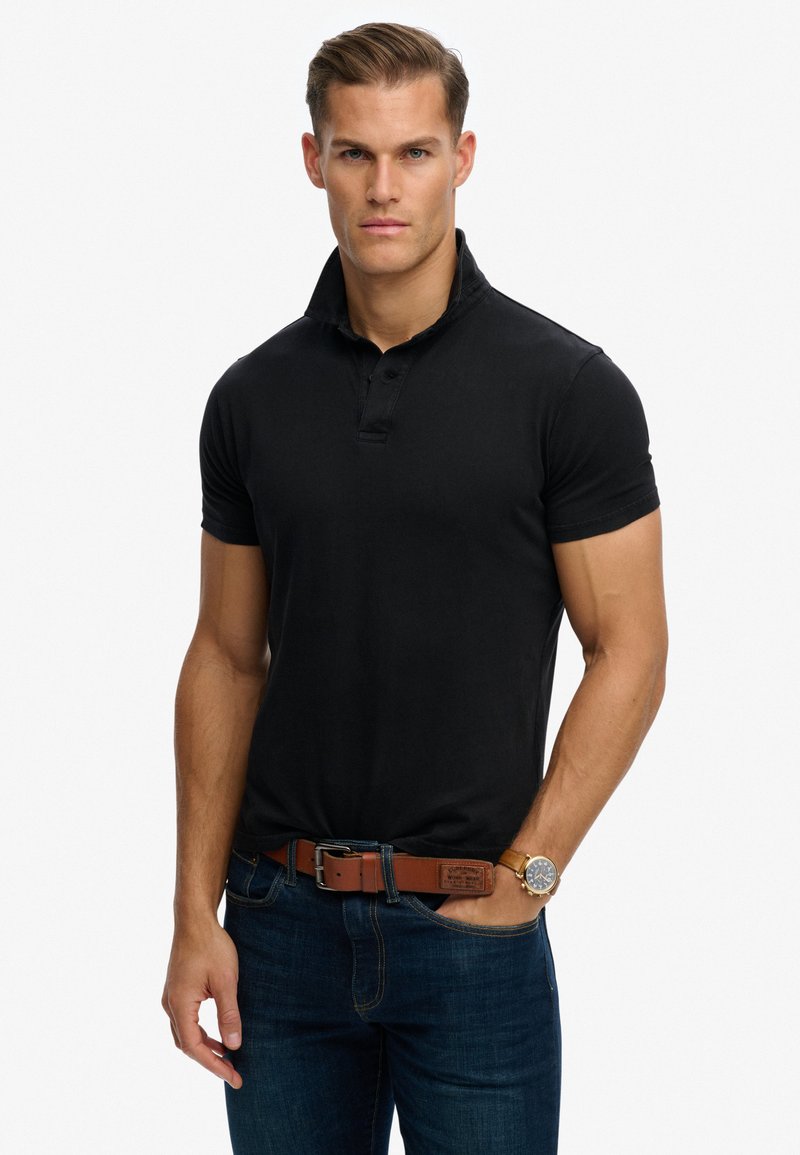 Polo-shirt noir avec un col court et une patte de boutonnage, porté avec un jean bleu et une ceinture en cuir marron. Design simple et en coton.