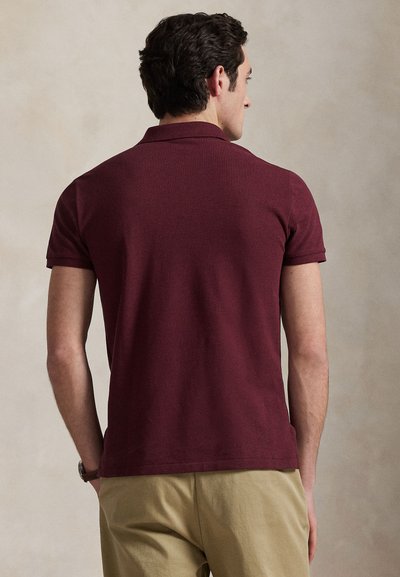 Homme aux cheveux bouclés foncés, portant un polo bordeaux à manches courtes et un pantalon beige, debout de dos sur un fond neutre.