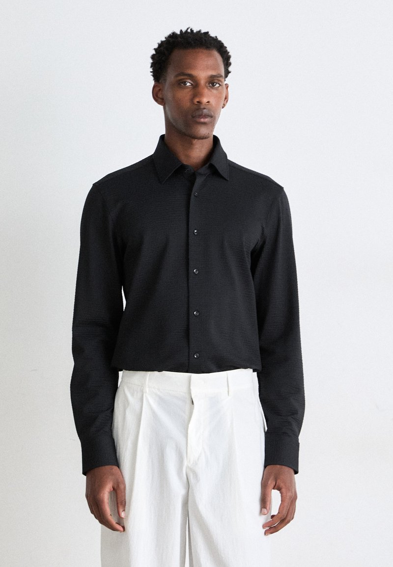 Camisa negra de manga larga con un tejido texturizado, frente abotonado y cuello puntiagudo, combinada con pantalones blancos con pliegues.