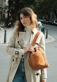 Brown suede drawstring bag with leather accents, rounded shape, soft texture, and visible hardware, carried on a shoulder strap.