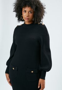 Schwarzes geripptes Strickkleid mit langen Puffärmeln, goldenen Knopfakzenten an den Schultern und zwei vorderen Taschen mit Klappenverschluss.