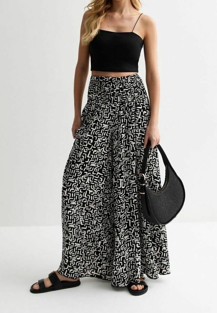 Femme portant un crop top noir à bretelles spaghetti, un pantalon large à motif noir et blanc, des sandales noires, et tenant un sac à main en forme de croissant noir.