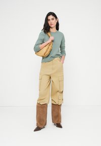 Πράσινο πουλόβερ με μακριά μανίκια, μπεζ cargo παντελόνι με πλαϊνές τσέπες, καφέ γούνινες λεπτομέρειες στους καρπούς και μουσταρδί τσάντα χειρός με κρόσσια.