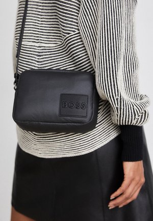 Sac bandoulière en cuir noir avec le logo « BOSS », porté sur un pull rayé noir et blanc et une jupe noire.