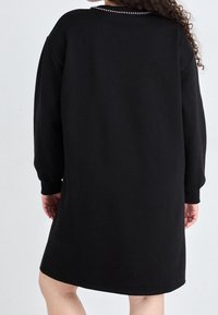 Robe sweat noire à manches longues, décolleté arrondi et léger éclat sur le tissu. Présente des embellissements discrets au niveau du col.