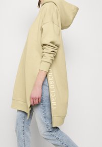 Sweat à capuche beige avec manches longues et fentes latérales. Présente un texte imprimé blanc le long de l'ourlet et une coupe décontractée. Associé à un jean bleu clair.