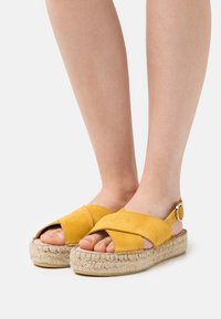 Yellow suede sandal with cross-over straps, open toes, and a jute platform sole. Adjustable ankle strap with metal buckle detail.