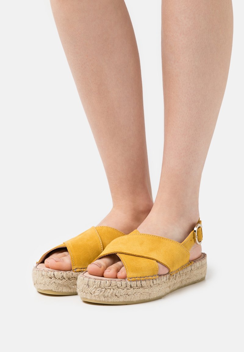 Yellow suede sandal with cross-over straps, open toes, and a jute platform sole. Adjustable ankle strap with metal buckle detail.