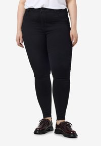 Personne portant un jean skinny noir, une chemise blanche et des chaussures à lacets bordeaux foncé, debout sur un fond blanc.