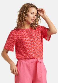 Rotes Kurzarmhemd mit einem weißen Blättermuster, kombiniert mit rosa Hosen, die einen Bindegürtel haben. Der Stoff wirkt leicht und strukturiert.