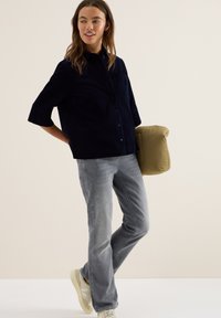 Marineblauwe corduroy shirt met een borstzak, gedragen met lichtgrijze jeans en beige sneakers, terwijl een klein, puffy olijfgroen kussen wordt vastgehouden.
