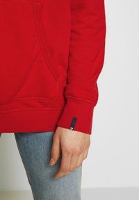 Main droite reposant à côté d'un jean bleu clair, portant un sweat-shirt rouge avec une poche avant et une petite étiquette en tissu noir sur le poignet.