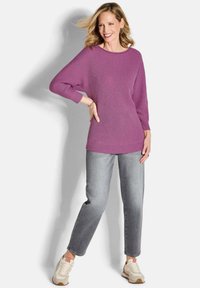Lächelnde Frau mit blonden Haaren trägt einen lila Strickpullover, graue Jeans und weiße Sneakers und steht mit der Hand in der Hüfte vor einem weißen Hintergrund.