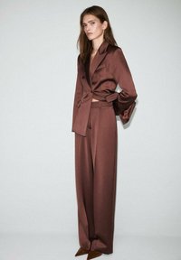 Brauner Satin-Zweiteiler, bestehend aus einem cropped, doppelt geknöpften Blazer und hochgeschnittenen, weiten Hosen. Spitz zulaufende braune Absätze runden den Look ab.