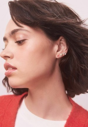 Drei Ohrringe in Herzform: einer in Rot, einer in Gold und einer in Silber, getragen am Ohr eines Models vor neutralem Hintergrund.