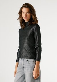 Μαύρο, εφαρμοστό μπλουζάκι με μακρύ μανίκι και ψηλό λαιμό. Κατασκευασμένο από μια υφή μεταλλικού υλικού που αντανακλά το φως. Συνδυασμένο με ανοιχτό γκρι τζιν.