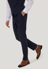 Granatowe spodnie garniturowe w połączeniu z białą koszulą z długim rękawem i brązowymi skórzanymi butami typu double monk strap. Wyraźne gładkie tekstury i dopasowany krój.