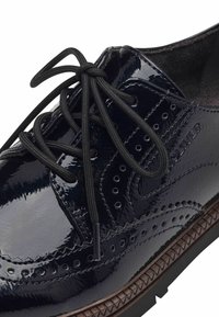 Chaussure en cuir verni noir avec un design à la pointe d'aile, comportant des perforations, des lacets noirs et une semelle brune texturée.