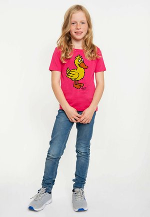 Rosa T-Shirt mit einem cartoonhaften gelben Entenmotiv. Das Design hat schwarze Umrisse. Kombiniert mit blauen Jeans und grauen Turnschuhen.