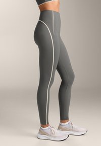 Vysoké šedé legíny so bielymi bočnými pruhmi, vyrobené z elastického materiálu. Skombinované s bielymi bežeckými topánkami pre dokonalý look.