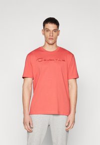 T-shirt en coton rouge à manches courtes et col rond, avec le logo "Champion" brodé sur la poitrine. Texture douce et lisse.