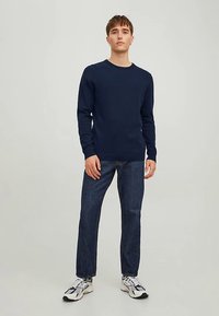 Navy blue long-sleeve shirt with a crew neck, paired with dark jeans and white sneakers, standing on a light gray background.
