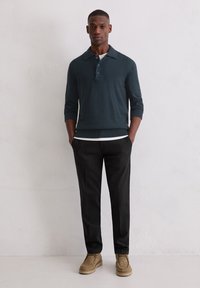 Ciemnozielony sweter z dzianiny z kołnierzykiem, zestawiony z czarnymi spodniami i jasnobrązowymi zamszowymi butami. Model stoi z rękami w kieszeniach.