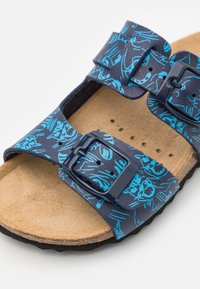 Sandal con patrón floral azul, dos correas ajustables, plantilla de corcho y suela de goma. Presenta una textura suave y hebillas resistentes.